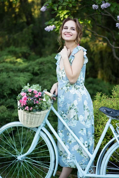 Retrato de una hermosa niña en el bosque, sosteniendo una bicicleta con una cesta de flores, detrás de los rayos del sol, un vestido de flores azules, paseo de verano —  Fotos de Stock