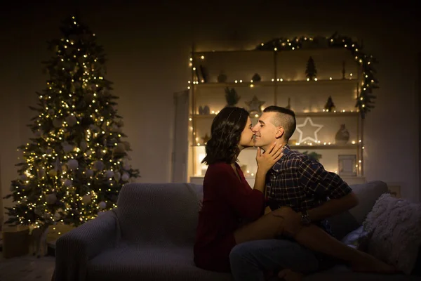 快乐，夫妻相爱在黑暗中，夜晚在沙发上，黑暗的背景，圣诞树上的灯。 平安夜 新年。 拥抱和亲吻。 迷人的，浪漫的夜晚和约会. — 图库照片