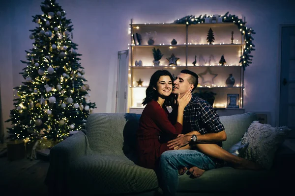 Feliz, casal apaixonado no escuro, noite no sofá, fundo escuro, luzes da árvore de natal. Noite de Natal. Ano Novo. abraço e beijo. atração, noite romântica e data . — Fotografia de Stock