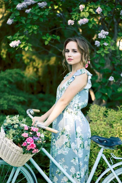 Retrato de uma menina bonita na floresta, segurando uma bicicleta com uma cesta de flores, atrás dos raios do sol, um vestido de flor azul, passeio de verão — Fotografia de Stock