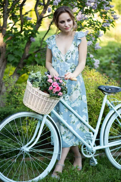 Portret pięknej dziewczyny w lesie, trzymając rower z koszem kwiatów, za promieniami słońca, niebieską kwiecistką sukienkę, letni spacer — Zdjęcie stockowe