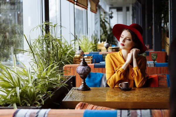 Fille dans un café avec une tasse de café et un chapeau.portrait de la jeune fille sensuelle portant un chapeau disquette et chemisier avec arc. Belle femme brune dans un café tenant une tasse de café — Photo