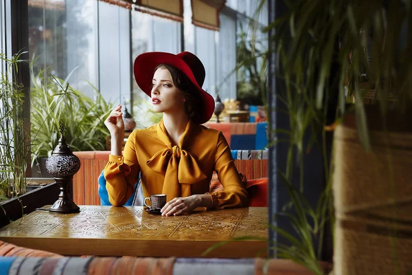 Fille dans un café avec une tasse de café et un chapeau.portrait de la jeune fille sensuelle portant un chapeau disquette et chemisier avec arc. Belle femme brune dans un café tenant une tasse de café — Photo