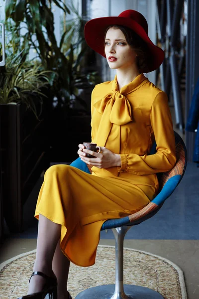 Dziewczyna w kawiarni z filiżanką kawy i kapelusikiem.Portret zmysłowej młodej dziewczyny w dyskietce kapelusz i bluzka z kokardą. Piękna brunetka w kawiarni trzyma filiżankę kawy — Zdjęcie stockowe
