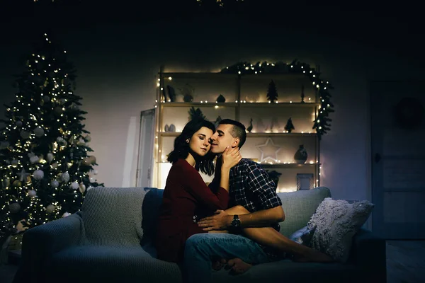 Feliz, casal apaixonado no escuro, noite no sofá, fundo escuro, luzes da árvore de natal. Noite de Natal. Ano Novo. abraço e beijo. atração, noite romântica e data . — Fotografia de Stock