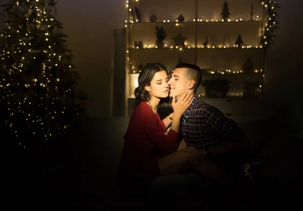 Feliz, casal apaixonado no escuro, noite no sofá, fundo escuro, luzes da árvore de natal. Noite de Natal. Ano Novo. abraço e beijo. atração, noite romântica e data . — Fotografia de Stock