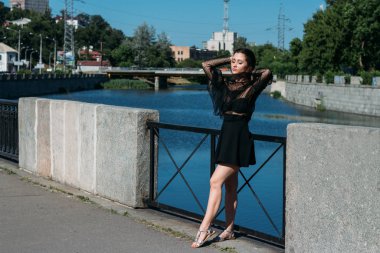 Güzel esmer kadın köprüde duruyor, nehrin yanında, şehirde. Siyah elbiseli bir kız yüzünü güneşe çevirir ve rüzgar onun üzerine eser.