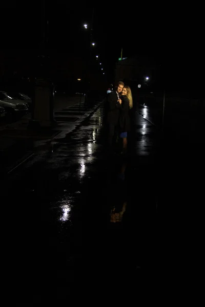 Ζευγάρι στην αγάπη αγκαλιάζει σε ένα σκοτεινό φόντο, νύχτα, βροχή, κτίρια στην πόλη, ένα πάρκο, χαμογελώντας, έλξη. Ο τύπος αγκαλιάζει το κορίτσι. Ημέρα του Αγίου Βαλεντίνου — Φωτογραφία Αρχείου