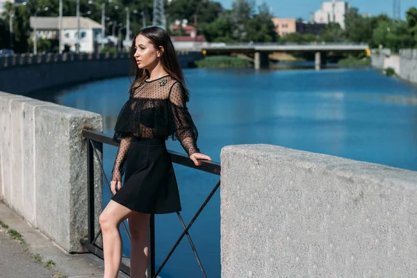 Красивая брюнетка стоит на мосту, возле реки, в городе. девушка в черном платье смотрит на солнце и ветер, который дует на нее — стоковое фото