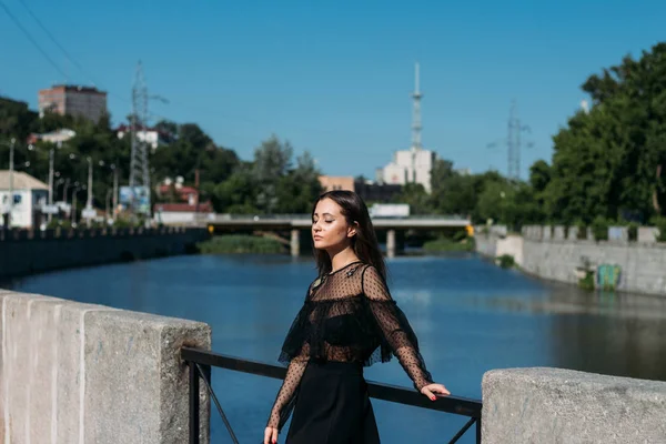 Beautiful brunette stands on the bridge, near the river, in the city. a girl in a black dress puts a face to the sun and the wind that blows on her — Stock Photo, Image