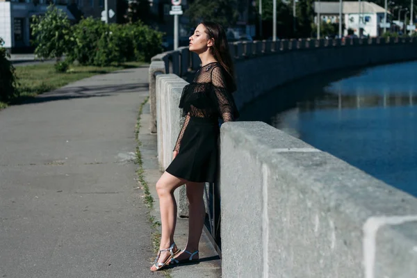 Hermosa morena se encuentra en el puente, cerca del río, en la ciudad. una chica en un vestido negro pone una cara al sol y el viento que sopla sobre ella —  Fotos de Stock