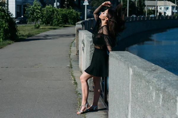 Красивая брюнетка стоит на мосту, возле реки, в городе. девушка в черном платье смотрит на солнце и ветер, который дует на нее — стоковое фото