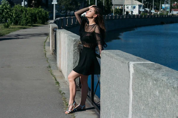 Hermosa morena se encuentra en el puente, cerca del río, en la ciudad. una chica en un vestido negro pone una cara al sol y el viento que sopla sobre ella —  Fotos de Stock