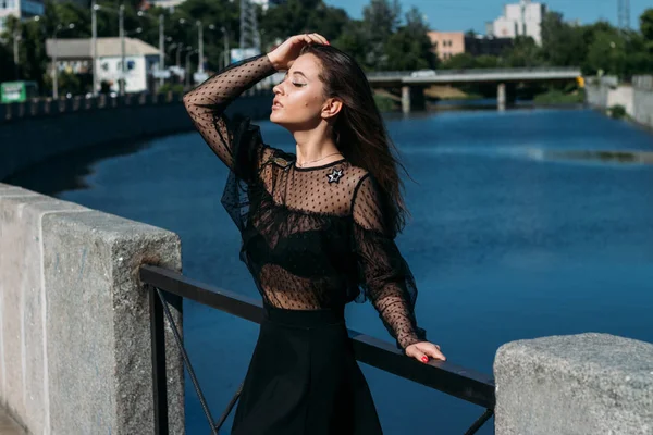 Gyönyörű barna áll a hídon, közel a folyóhoz, a városban. Egy fekete ruhás lány arcot vág a naphoz, és a szél fúj rá. — Stock Fotó