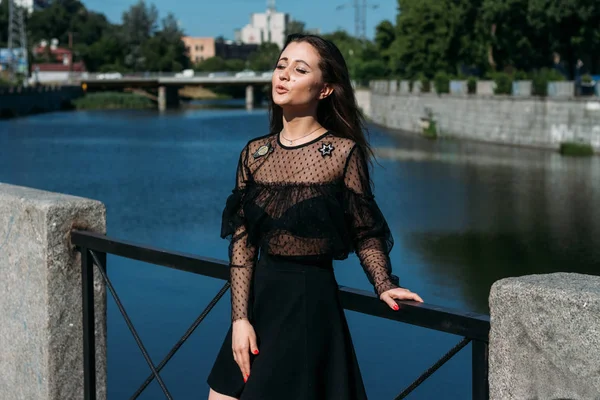 Belle brune se dresse sur le pont, près de la rivière, dans la ville. une fille en robe noire met un visage au soleil et le vent qui souffle sur elle — Photo