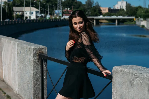 Bella bruna si trova sul ponte, vicino al fiume, in città. una ragazza in abito nero mette un volto al sole e il vento che soffia su di lei — Foto Stock