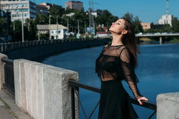 Красива брюнетка стоїть на мосту, біля річки, в місті. дівчина в чорній сукні ставить обличчя до сонця і вітру, який дме на неї — стокове фото