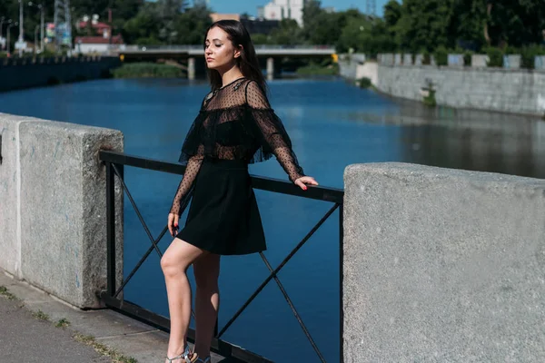 Bella bruna si trova sul ponte, vicino al fiume, in città. una ragazza in abito nero mette un volto al sole e il vento che soffia su di lei — Foto Stock