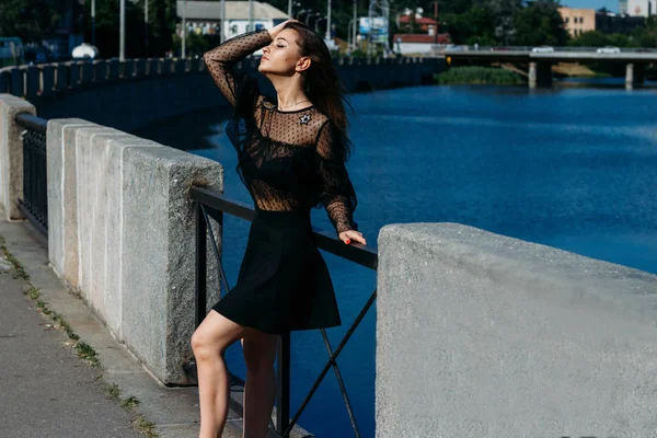 Belle brune se dresse sur le pont, près de la rivière, dans la ville. une fille en robe noire met un visage au soleil et le vent qui souffle sur elle — Photo