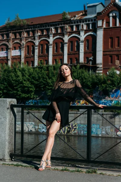 Bella bruna si trova sul ponte, vicino al fiume, in città. una ragazza in abito nero mette un volto al sole e il vento che soffia su di lei — Foto Stock