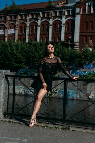 Όμορφη μελαχρινή στέκεται στη γέφυρα, κοντά στο ποτάμι, στην πόλη. Ένα κορίτσι με μαύρο φόρεμα βάζει ένα πρόσωπο στον ήλιο και τον άνεμο που φυσάει πάνω της — Φωτογραφία Αρχείου