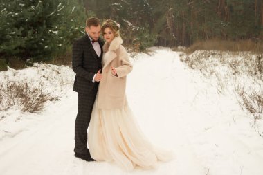 Çift sarılıyor ve öpücüğe yakın, el ele tutuşuyor, ellerinde bir buket çiçek. Kelebekli bir takım elbise ve elbiseli bir ceket. Gelin ve damat