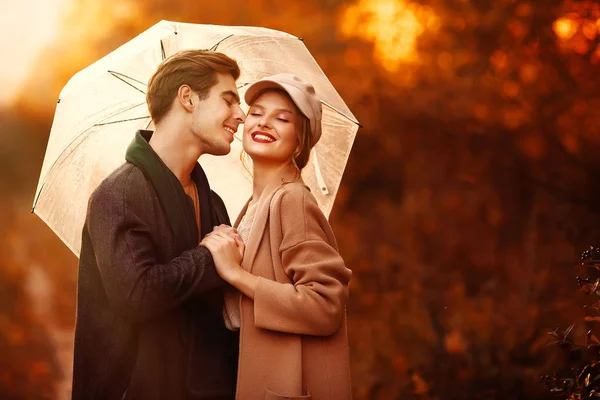 Happy, couple in love hugs and smiles on the street, autumn, green scarf and cap, man and woman on a date, valentines day. walk in the park under an umbrella in the rays of the setting sun — Stock Photo, Image