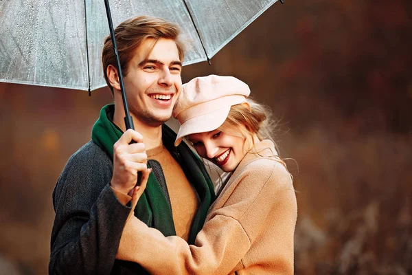 Feliz, casal no amor abraços e sorrisos na rua, outono, lenço verde e boné, homem e mulher em um encontro. caminhar no parque sob um guarda-chuva nos raios do sol poente — Fotografia de Stock