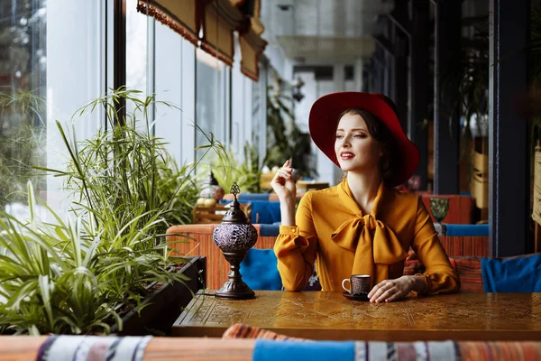 Fille dans un café avec une tasse de café et un chapeau.portrait de la jeune fille sensuelle portant un chapeau disquette et chemisier avec arc. Belle femme brune dans un café tenant une tasse de café — Photo