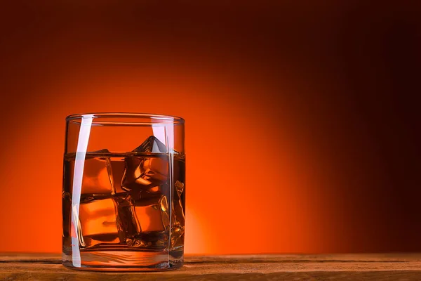 A glass of whiskey or cognac and ice cubes, close-up on a wooden table. Bright orange brown luminous background. Space for lettering, text, and logo. Layout for advertising. Brutal view.