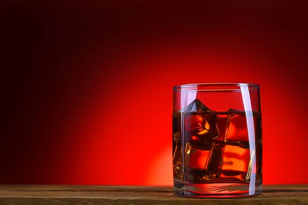 A glass of whiskey or cognac and ice cubes close-up on a wooden table. Bright red glowing background. Space for lettering, text, and logo. Layout for advertising.