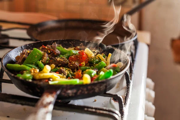 Red meat and colorful vegetables cooking in an old dirty fry pan, steam rising in a bright sunlight.