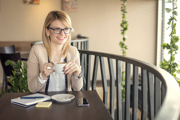 Young smiling business woman sitting in coffee shop, drinking coffee and thinking about something