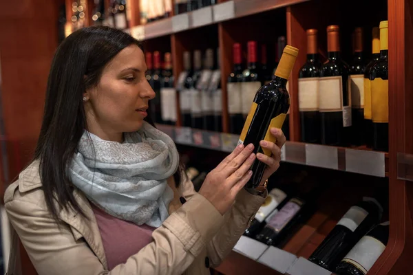 Portrait of happy young woman holding wine bottle in hands and smiling while standing in liquor store. Happy young woman choosing and buying wine in market. Sale, shopping, consumerism and people concept.