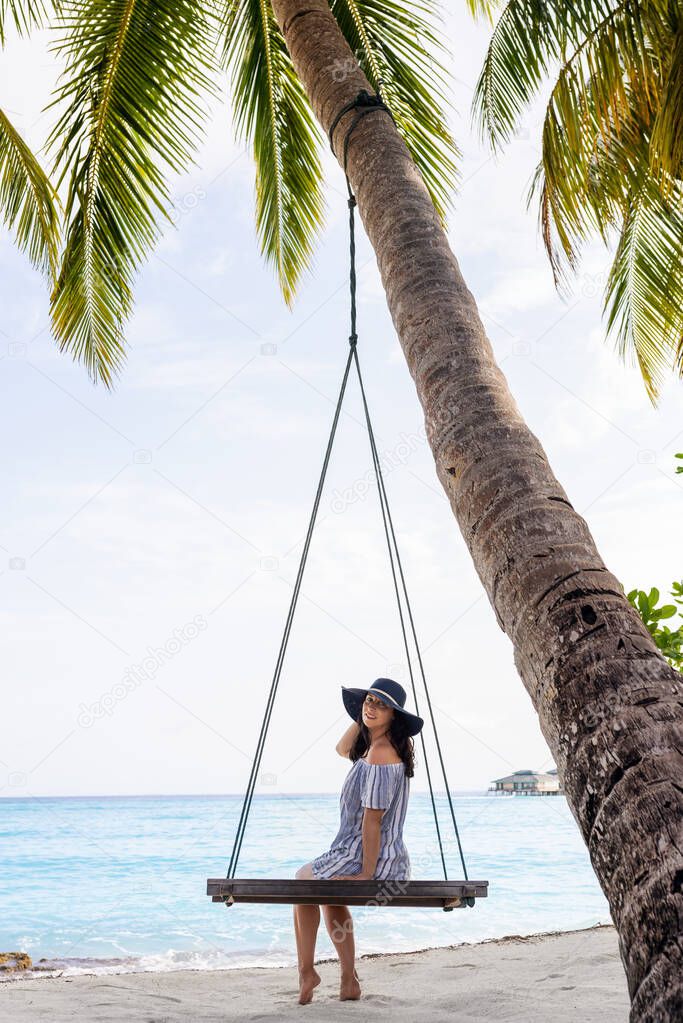 Beautiful fit girl on travel vacation. Portrait of smiling and attractive young woman at sea. Young beautiful woman relaxing in swing hanging on coconut palm at exotic beach.