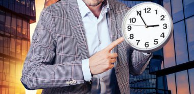 Takım elbiseli bir adam bir gökdelenin arka planındaki saate işaret ediyor. Zaman, iş, başarı kavramı..