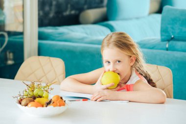Masada elinde defterle ödevini yapan küçük bir kız vardı ve taze bir elma yemeye karar verdi..
