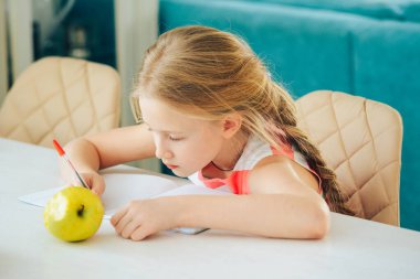 Masada küçük bir kız elinde defterle ödev yapıyor ve taze bir elma yiyor..