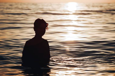 Kısa saçlı bir kızın silueti güneşin doğuşunun arka planında suyun içinde duruyor..