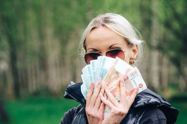 blonde girl made a fan out of a pack of rubles and hid her face bragging about money. clipart