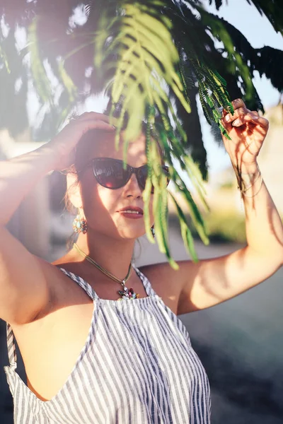 晴れた日に木の枝の後ろに隠れているサングラスの魅力的な女の子の肖像画 海で休暇中の熱帯の都市の観光スポットを歩く — ストック写真