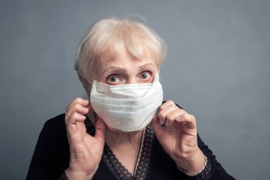 Gri arka planda, tıbbi maskeli yaşlı bir kadın. Kamera gribi salgınına, toza alerjiye ve virüse karşı korunmaya bakan bir yetişkinin stüdyo portresi.