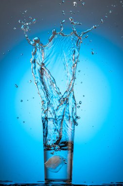 Bir bardakta büyük bir sıçrama ve akıntı. Buz küpünden mavi bir arka planda temiz su var.