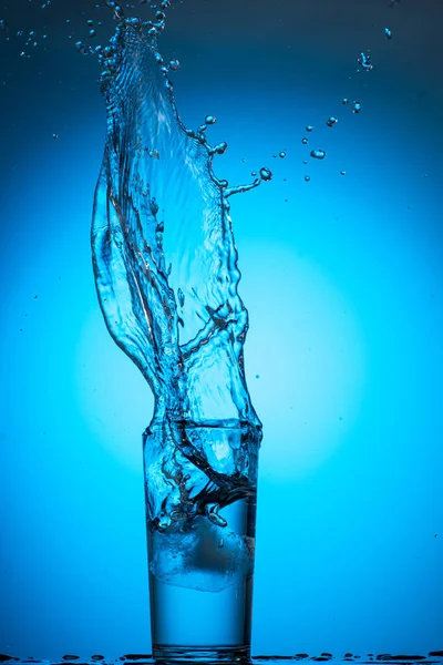 大的水花和溪流在一个玻璃杯中 在蓝色的青色背景上 从冰块中清澈的水 — 图库照片