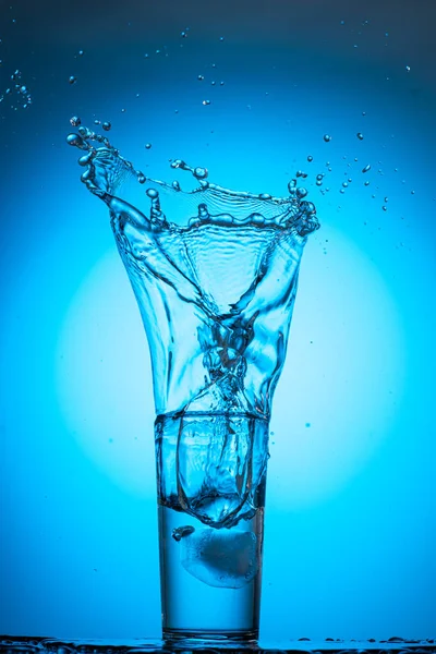 大的水花和溪流在一个玻璃杯中 在蓝色的青色背景上 从冰块中清澈的水 — 图库照片