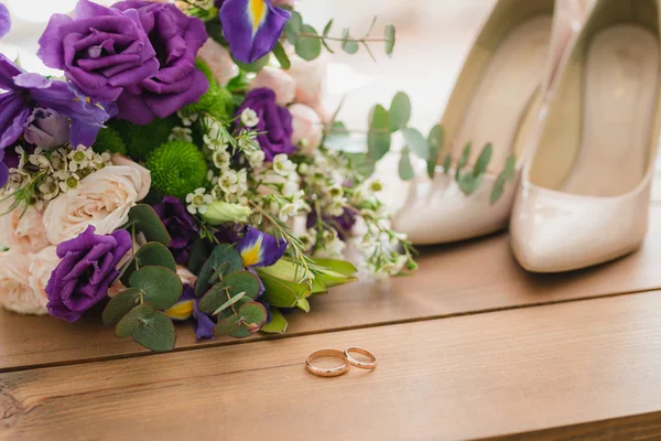 Sobre una mesa de madera un ramo de bodas con hojas verdes, flores púrpuras y blancas, anillos de boda — Foto de Stock