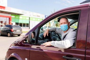 Koruyucu maskeli bir adam araba kullanıyor. Koruyucu maskeli bir adam araba sürerken eczaneye gider. Gri kazaklı bir adam arabada oturuyor ve koronavirüs maskesi takıyor..