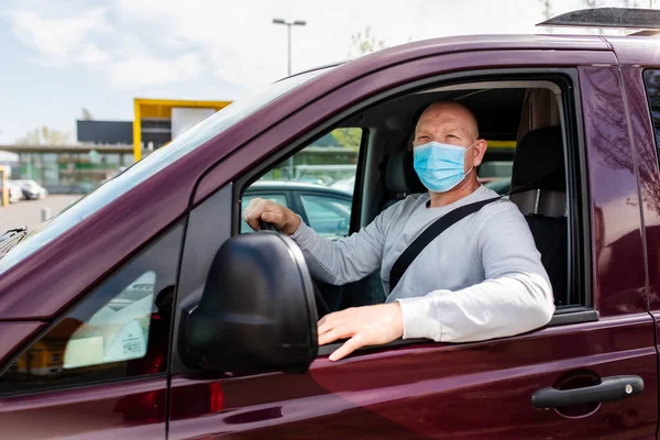 A man in a protective mask driving a car. A man in a protective mask driving a car goes to the pharmacy. A man in a gray sweater is sitting in a car and wearing a mask from a coronavirus.