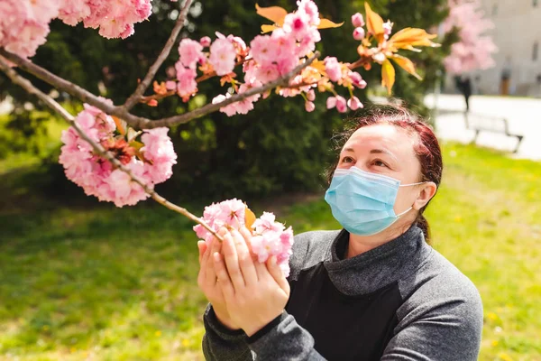 Parktaki kız koruyucu maskeli bir sakura çiçeğini kokluyor. Sakura parkında bir kadın yürüyor..