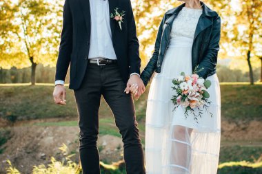 Gelin ve damat, güzel bir buketin ellerinde el ele tutuşuyorlar. Çift gün batımında çölde duruyor. Gelin ve damat gün batımında durup el ele tutuşuyorlar. Gelinlik ve siyah ceketli gelin..